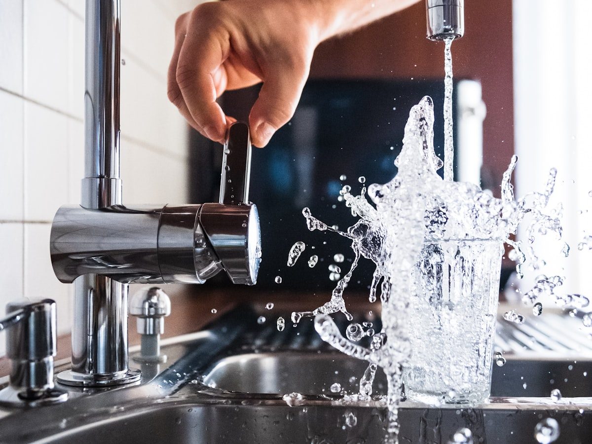 Water running hard from a kitchen faucet and splashing out of a glass into a stainless steel sink
